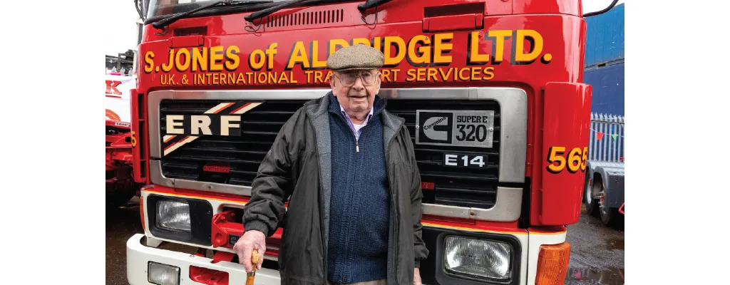 An elderly man wearing a flat cap, glasses, and a dark coat stands in front of a red ERF truck belonging to "S. Jones of Aldridge Ltd." He is holding a walking stick and has a slight smile on his face. The truck has bold yellow lettering and a Cummins "Super 320" badge.