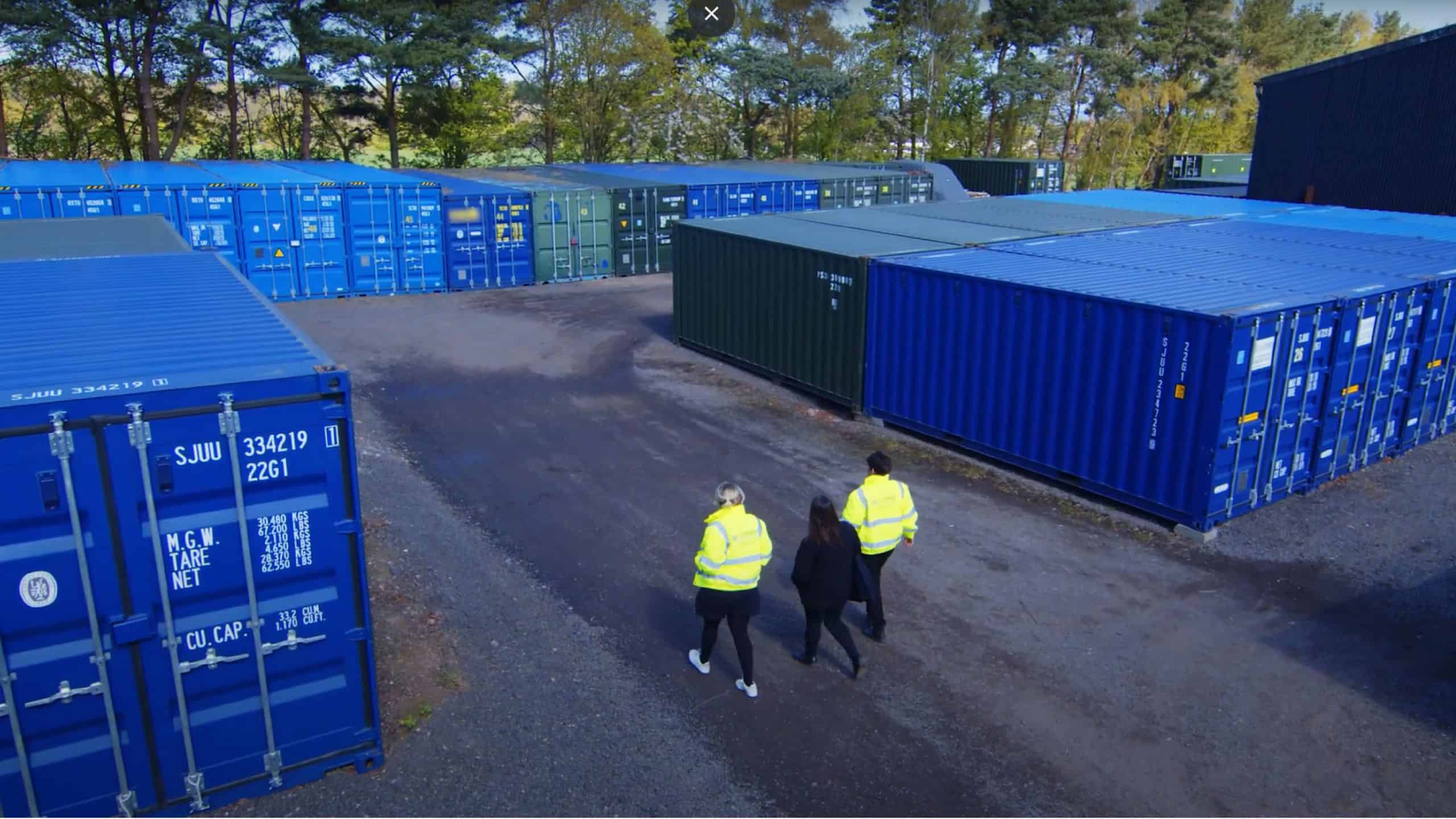 An outdoor self-storage facility with multiple shipping container units in blue and green, neatly arranged on a paved lot. Three people, two wearing high-visibility yellow jackets and one in black clothing, walk along a pathway through the storage area. The facility is surrounded by trees and greenery, with a mix of open and closed containers visible. The sky is clear, and the setting appears to be in a semi-rural or industrial location.