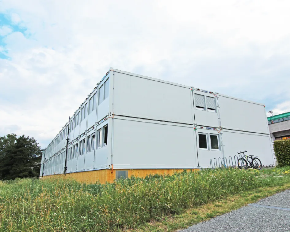 A large, two-story modular building made of white container units, situated on a grassy area with a gravel path leading up to it. A bicycle is parked near a metal rack beside the structure. The sky is blue with scattered clouds, and a nearby building under construction is visible in the background.