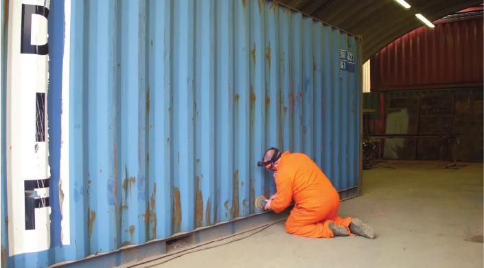 A worker in bright orange protective clothing and safety goggles is kneeling and using a handheld tool to grind or clean the rusted bottom edge of a large blue shipping container inside a workshop with arched roofing.