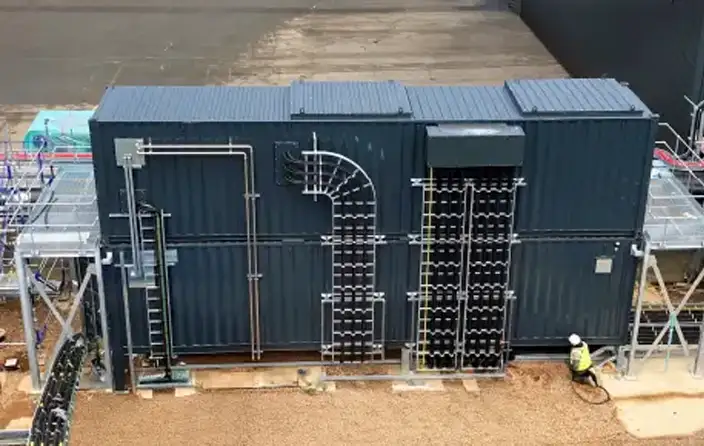 A dark-coloured modified shipping container for Cummins with extensive piping and external infrastructure, part of an industrial generator setup, with a worker in a high-visibility vest standing nearby.
