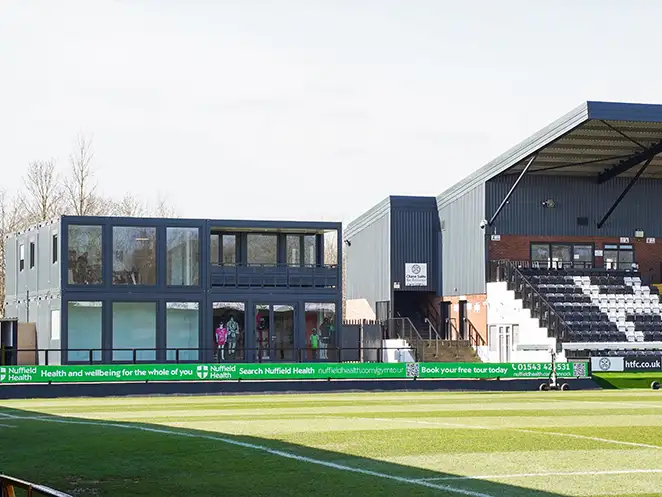 Exterior view of modular office units by S Jones Containers at Hednesford Town Football Club, positioned beside the football pitch with large windows, balcony, and integrated with the main stadium structure.