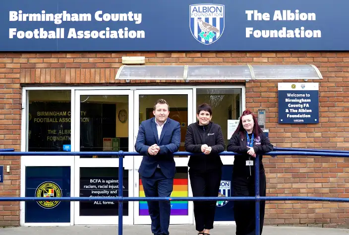 Three people stand smiling outside the Birmingham County Football Association and The Albion Foundation office, which has a blue sign with logos above the entrance and a rainbow flag displayed in the window.