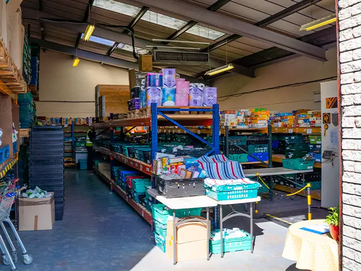 Interior of a warehouse with metal shelving units stocked with household goods such as toilet paper, cleaning products, and boxed items. Plastic crates are organized on the shelves, and a table at the front holds folded textiles and packaged items. Fluorescent lights and natural light from skylights illuminate the space.
