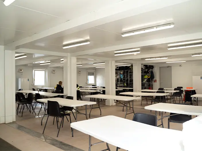 nterior of a modular building set up as a canteen with white tables, black chairs, fluorescent lighting, and a worker sitting at one of the tables